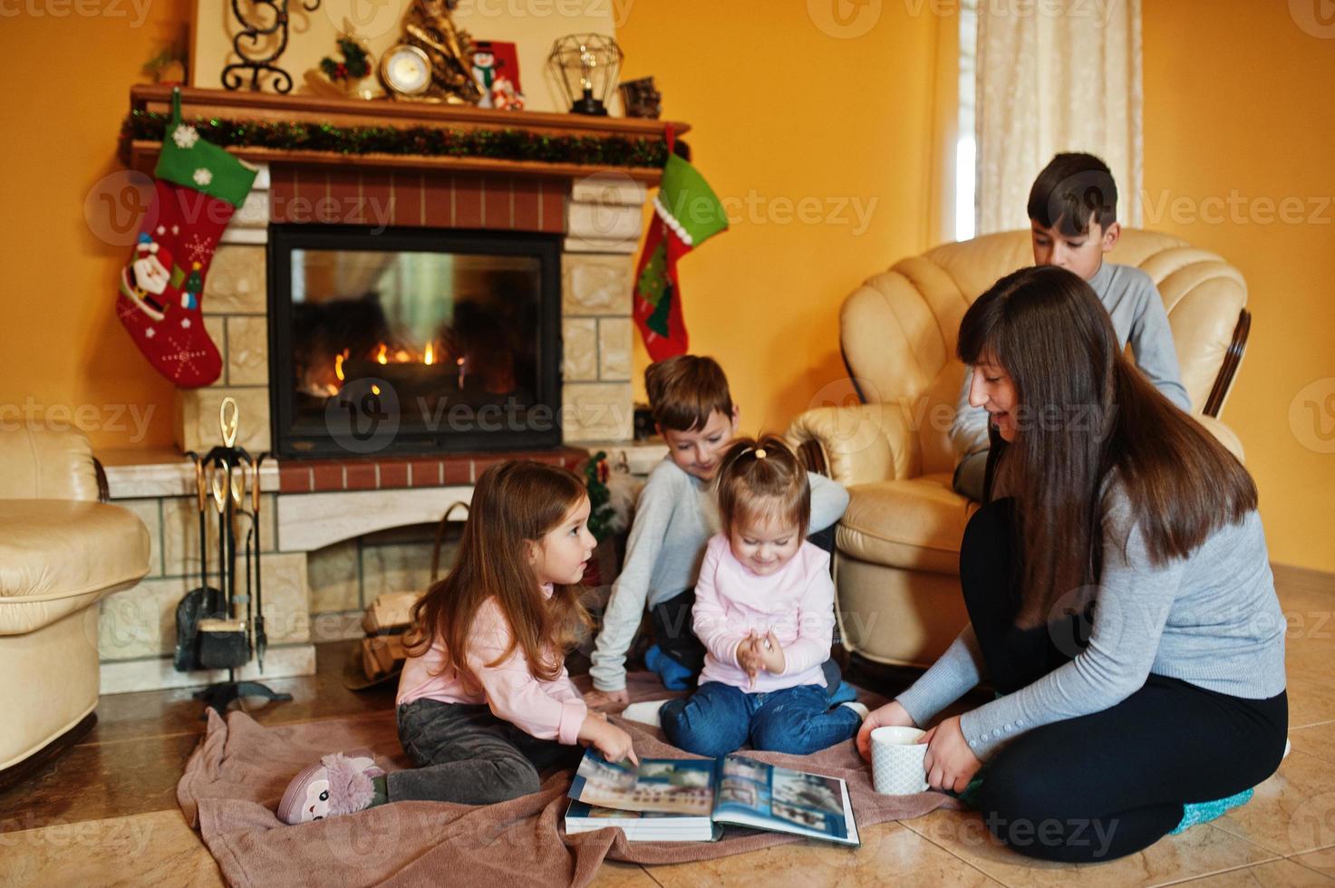 Happy young large family by a fireplace in warm living room on winter day. Mother with four kids at home read book. photo