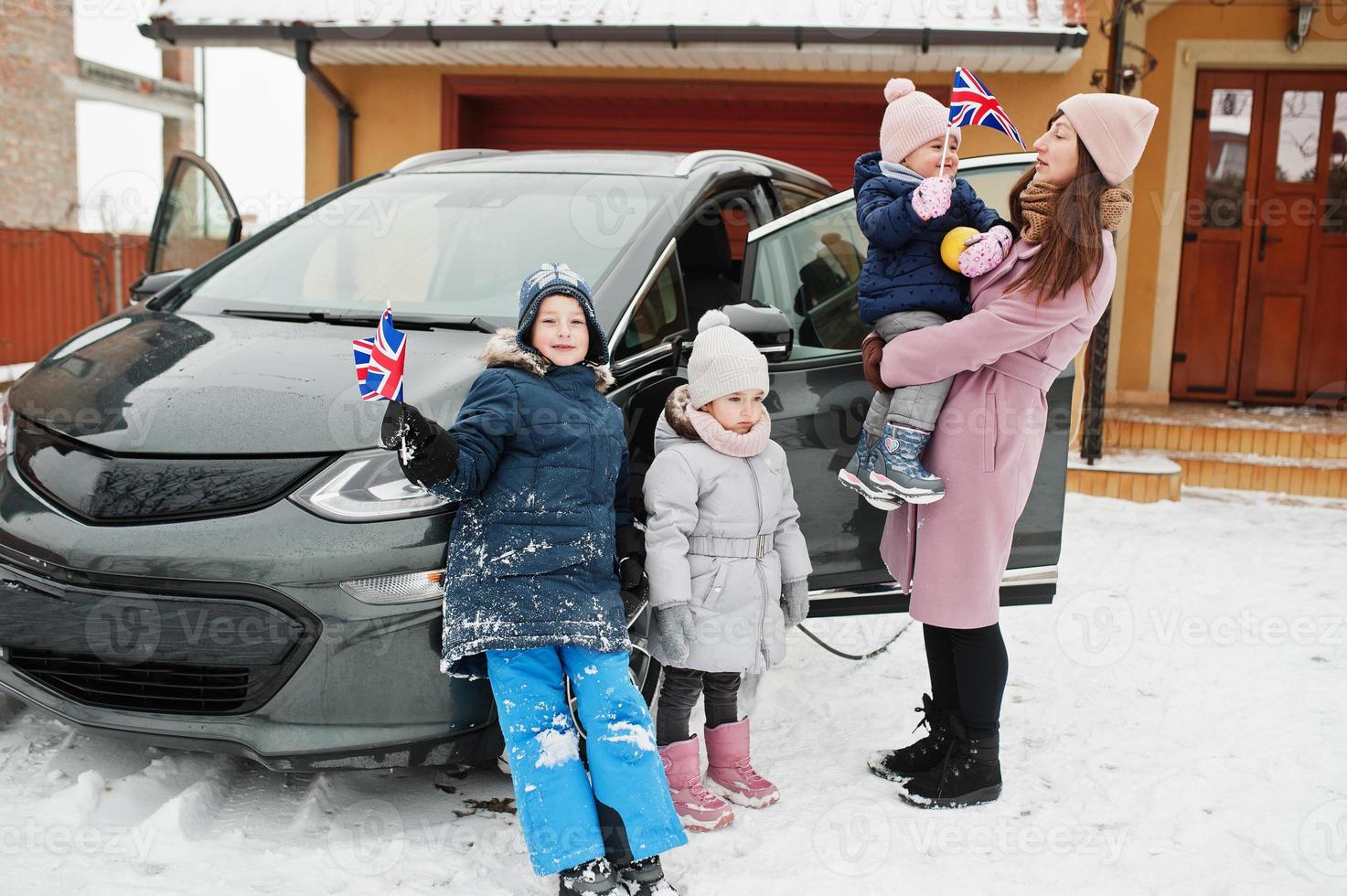 Young british mother with kids hold Great Britain flags and charging electric car in the yard of her house at winter. photo