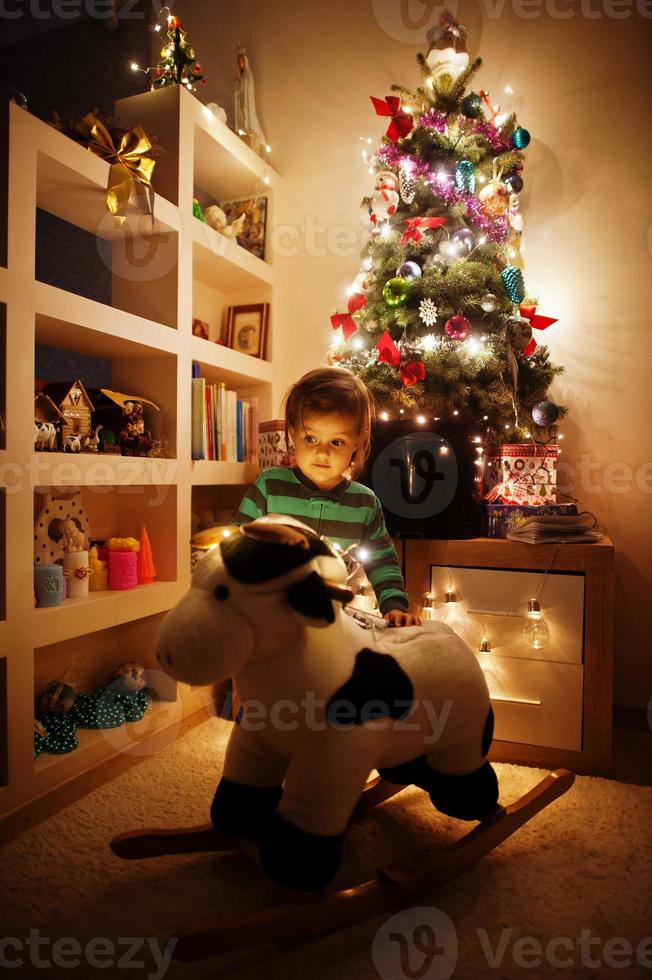 Baby girl on rocking cow toy against Christmas tree with shining garlands on evening home. photo