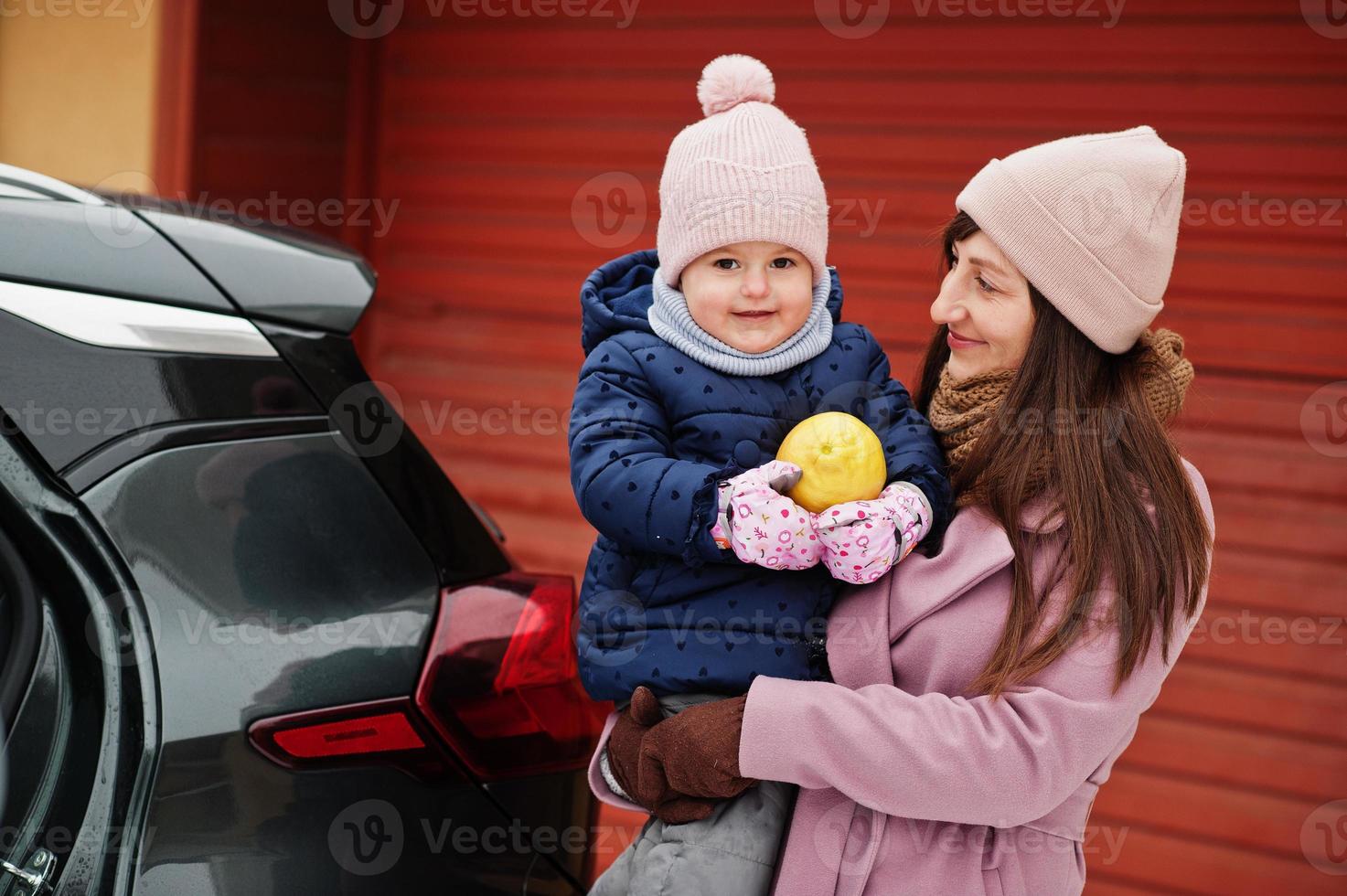 Young mother with baby girl on hands against car, hold lemon at winter. photo