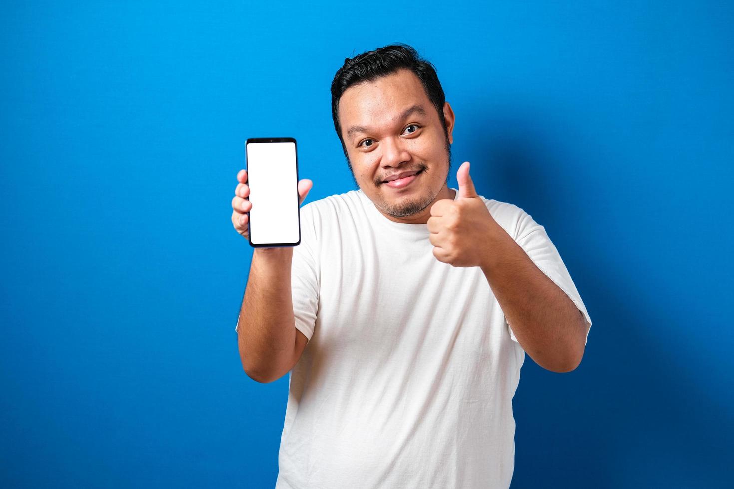 Pleased happy young fat asian guy in white t-shirt smiling to camera while holding smartphone pointing at cellphone screen photo