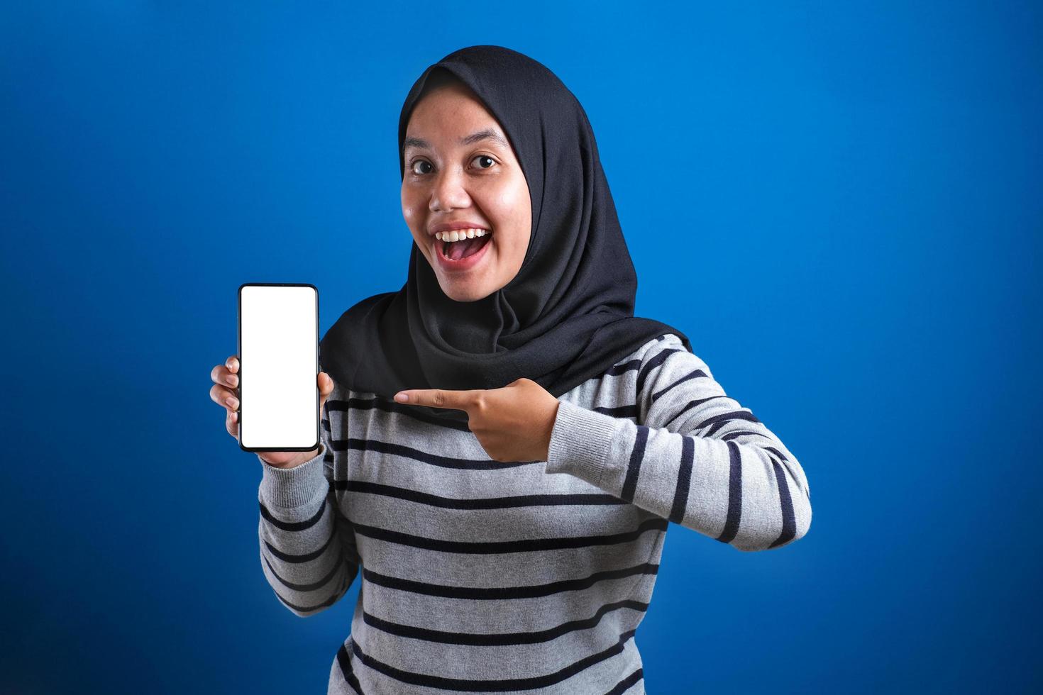 Portrait of Asian muslim woman looking at camera smiling, showing and pointing her smart phone, photo