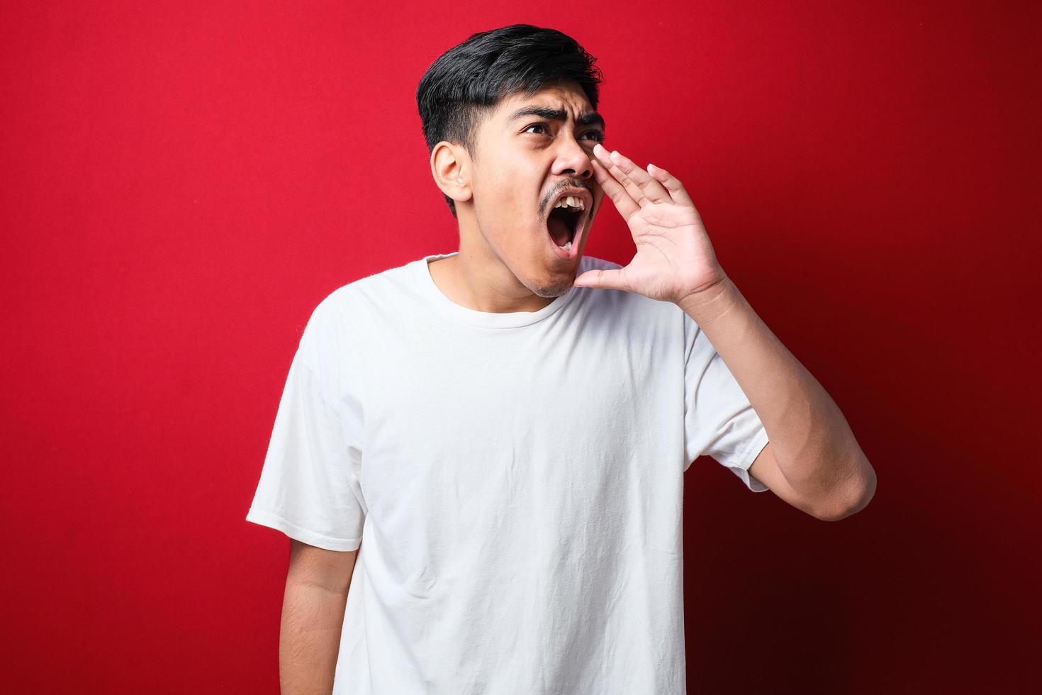 joven asiático con camiseta blanca gritando y gritando fuerte a un lado con la mano en la boca foto