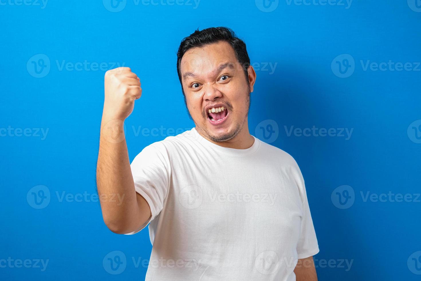 Portrait of a funny fat Asian man in white t-shirt smiling and dancing happily, joyful expressing celebrating good news victory winning success gesture against blue background photo