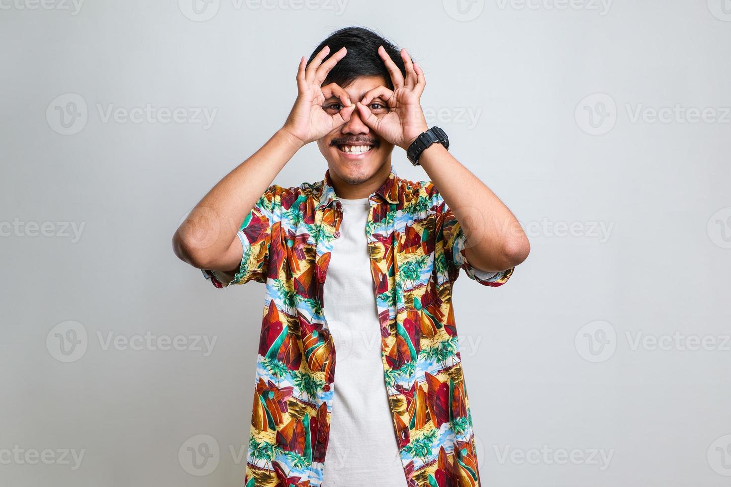 Man wearing casual shirt doing ok gesture like binoculars sticking tongue out, eyes looking through fingers photo