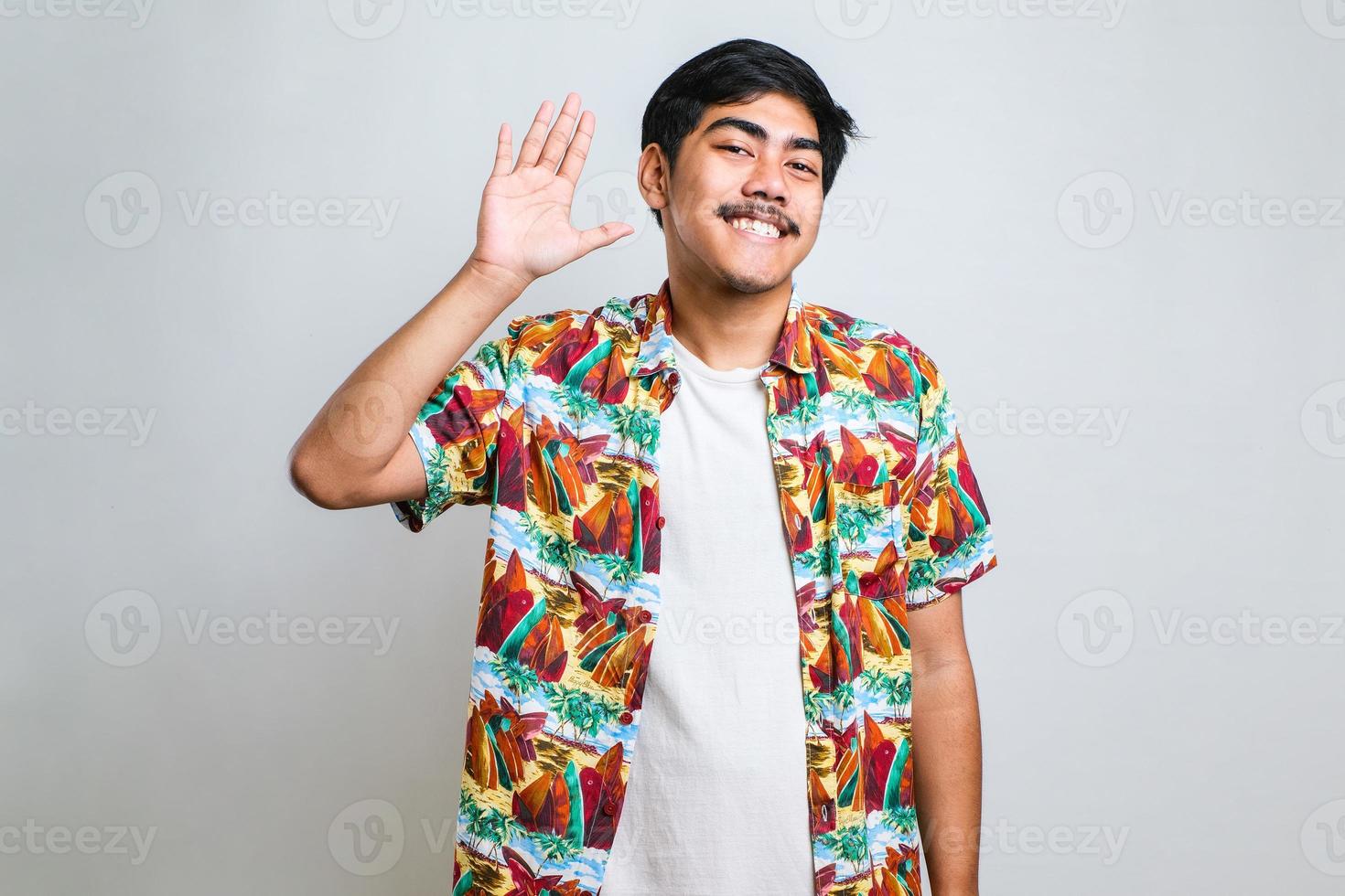 Man wearing white tshirt standing over white background Waiving saying hello happy and smiling, friendly welcome gesture photo
