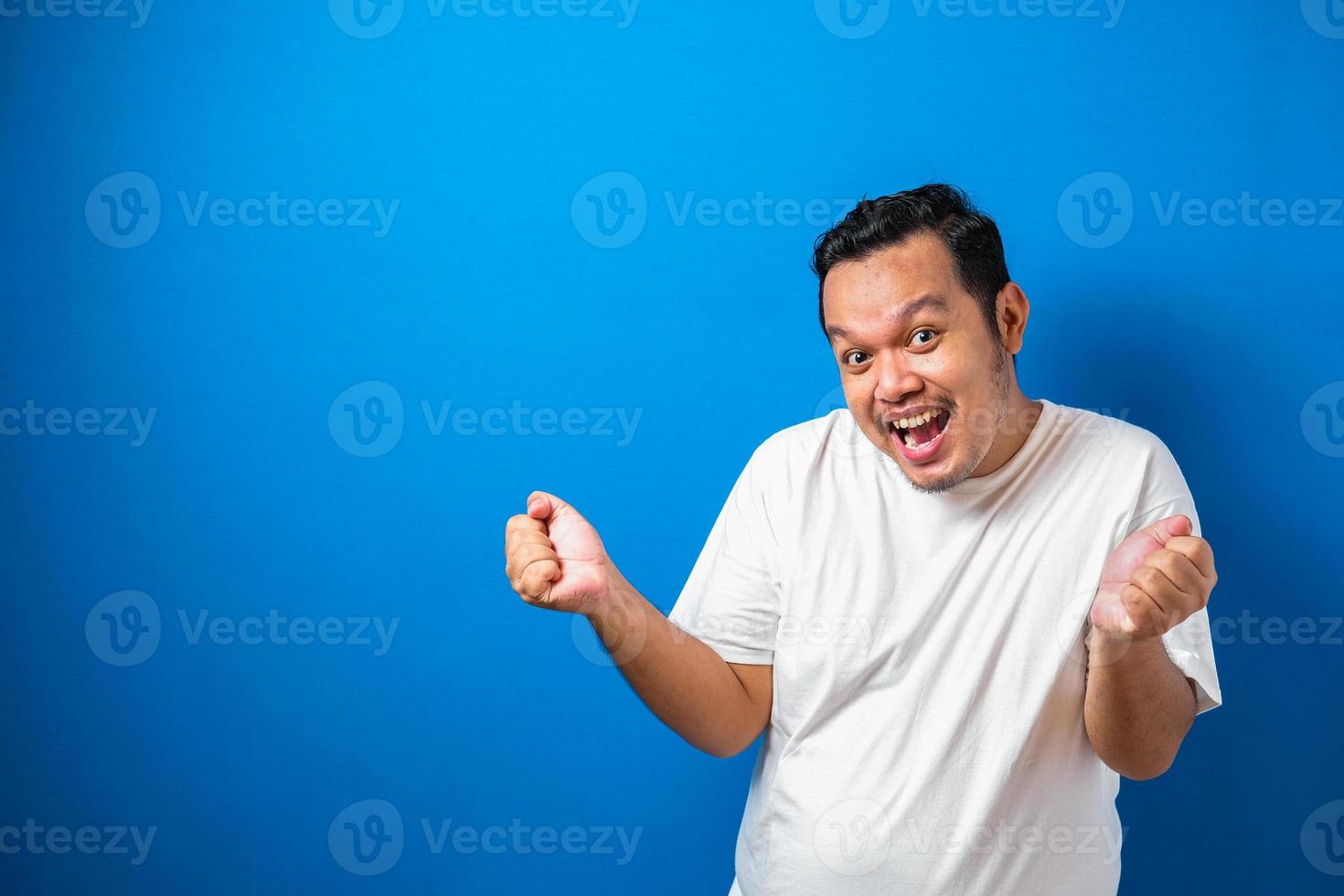 Portrait of a funny fat Asian man in white t-shirt smiling and dancing happily, joyful expressing celebrating good news victory winning success gesture against blue background photo