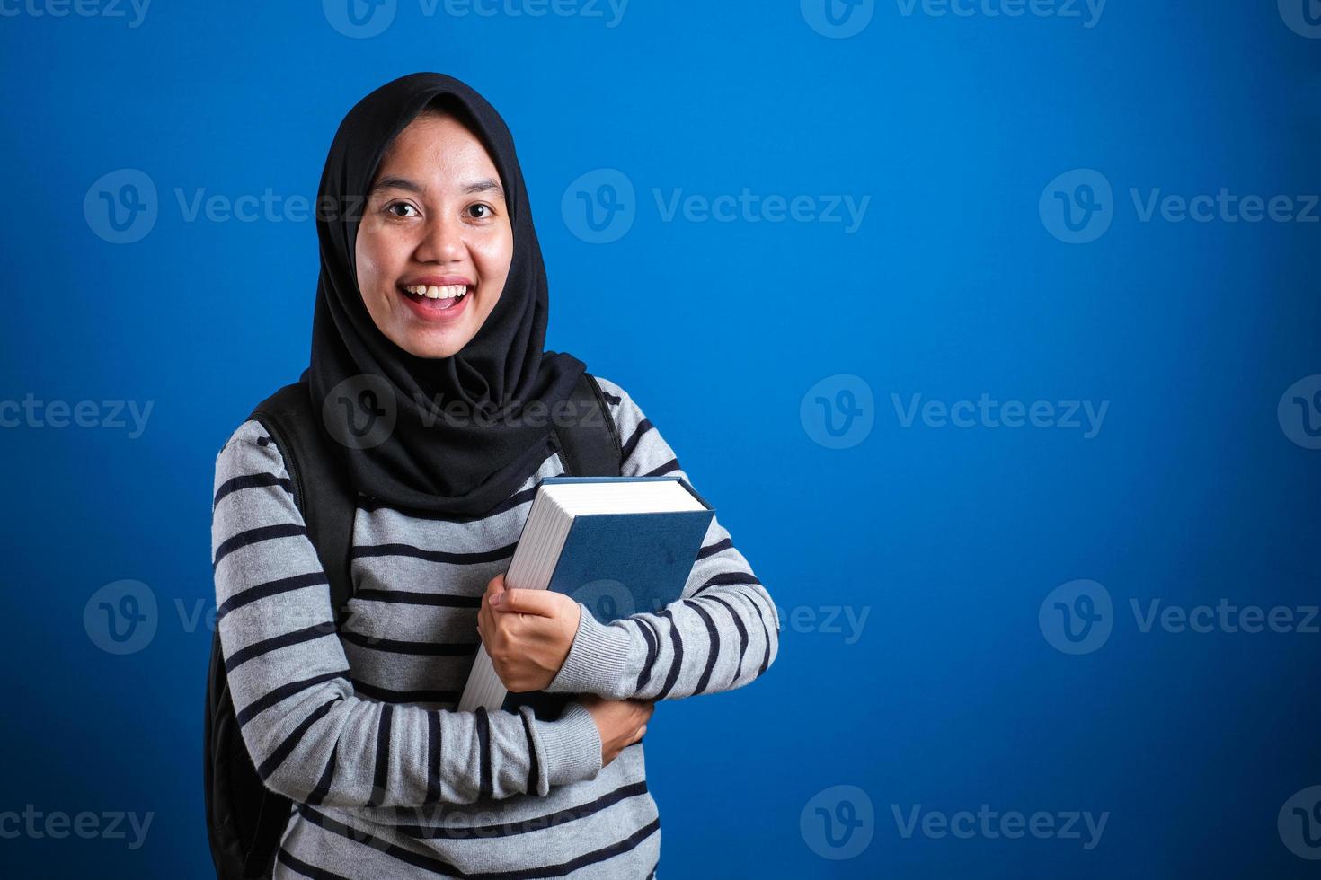 estudiante universitaria musulmana asiática usando hijab sonriendo amigable sosteniendo un libro grande foto