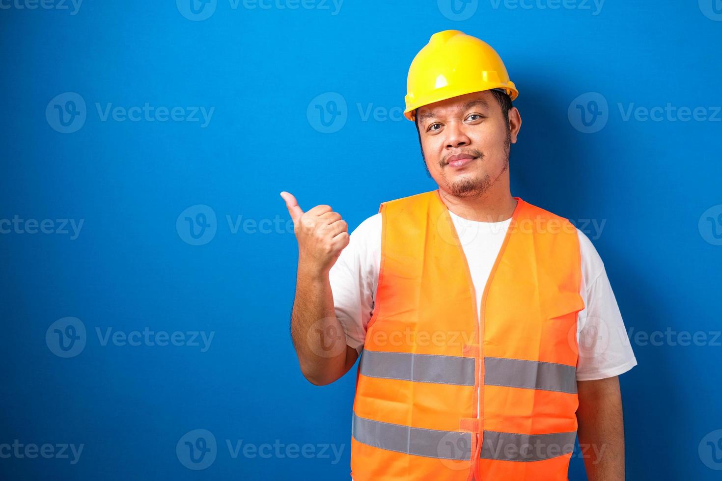 Young fat asian man over blue background wearing contractor uniform and safety helmet pointing and showing with his finger photo