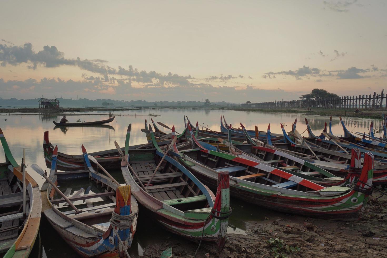 botes en la orilla del lago con un hombre sentado en un bote en el lago con el fondo del puente u-bein, mandalay, myanmar a la hora del amanecer foto