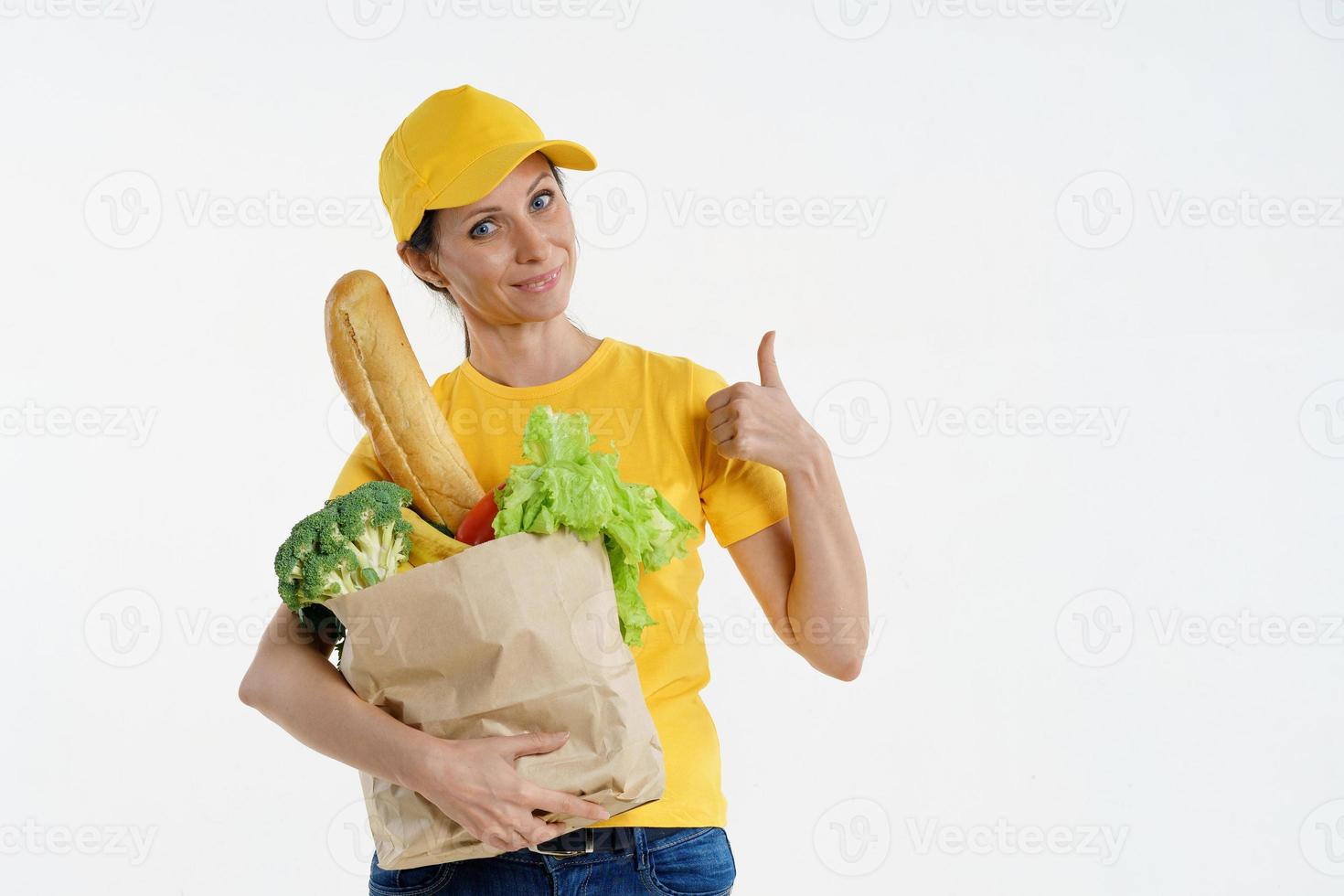 Delivery woman in yellow holding grocery bag and giving thumbs up, on white background photo