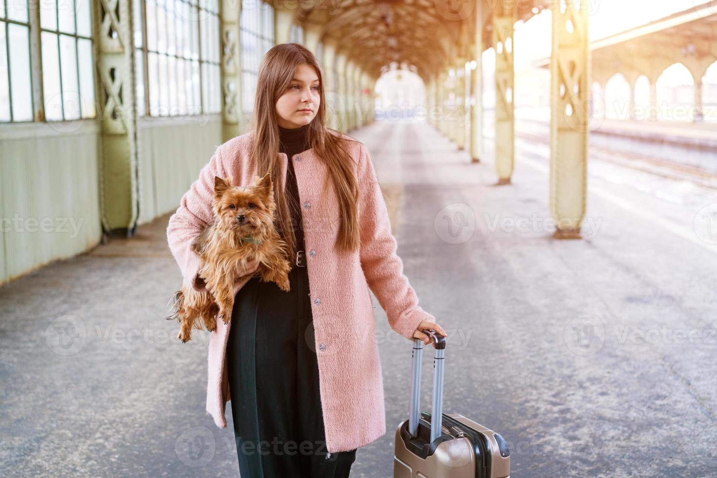 Travel concept. At station, young tourist with dog goes and drags suitcase photo