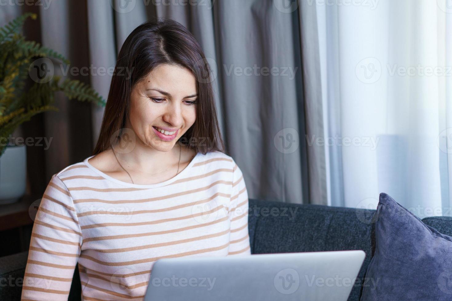 Smiling caucasian girl sitting on sofa at home, working on modern portable photo