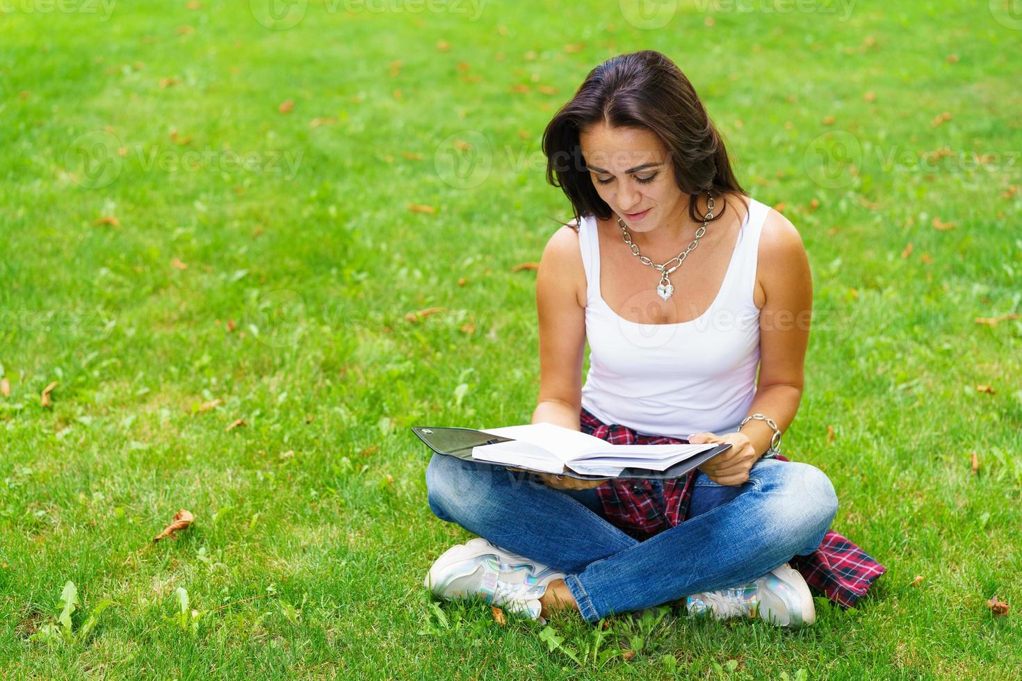 Smiling female student sitting on grass with notepad in hand, preparing photo