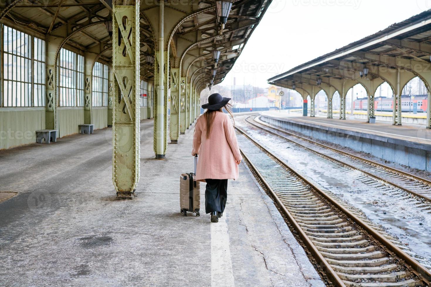 mujer con abrigo rosa camina con maleta en una plataforma vacía con la espalda foto