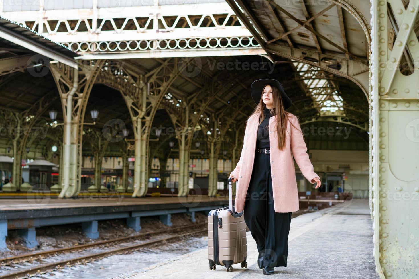 Beautiful young casual tourist woman with a suitcase in a pink coat waiting photo