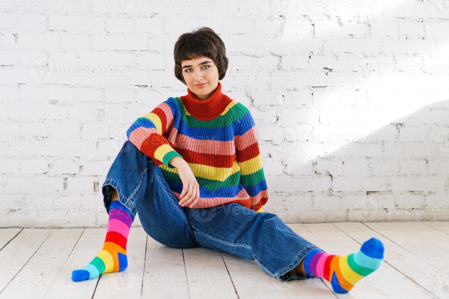 Cute cheerful young girl in colorful sweater, sitting on floor against photo