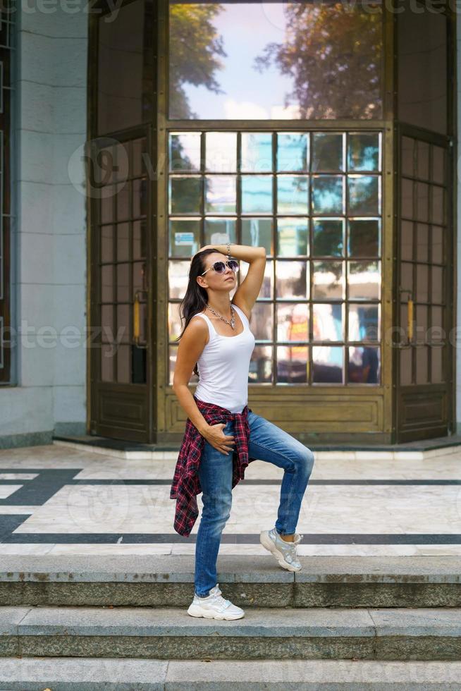 linda mujer con gafas de sol posa en los escalones de la calle cerca del edificio foto