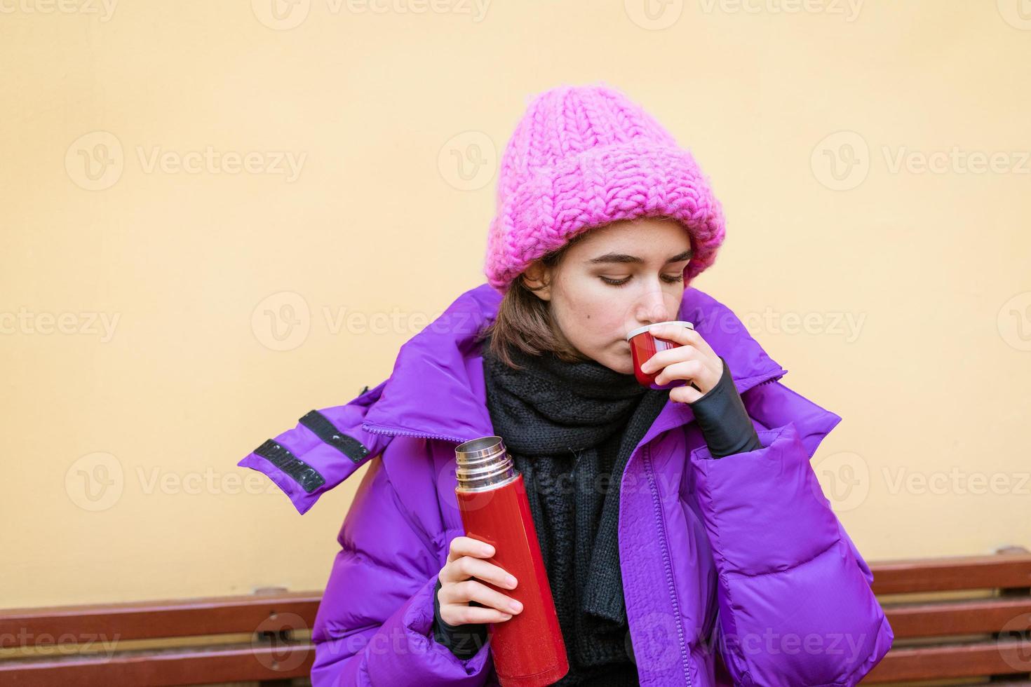 Young cute girl with dark hair in pink down jacket sits on wooden bench photo