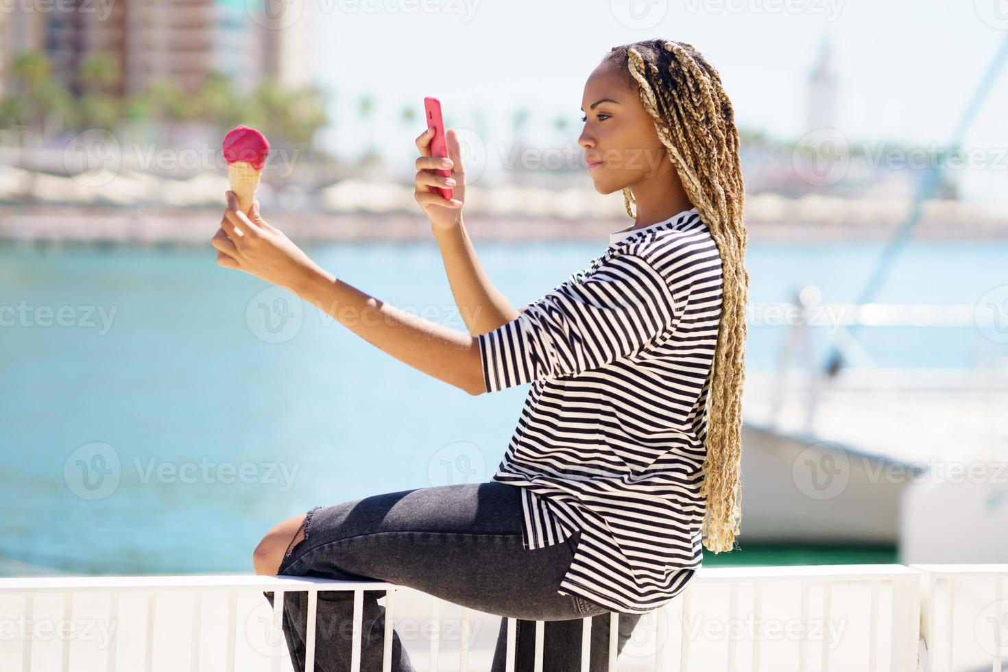 chica negra fotografiando con su smartphone un helado de fresa que está comiendo cerca del mar foto