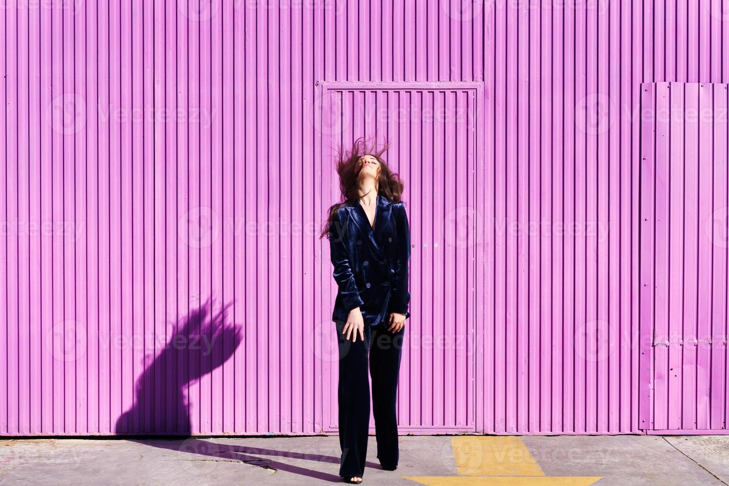 Woman wearing blue suit posing near pink shutter, moving her hair. photo