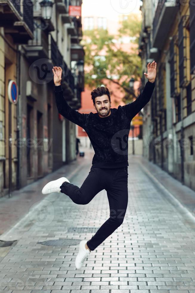Young bearded man jumping in urban background with open arms wearing casual clothes. photo