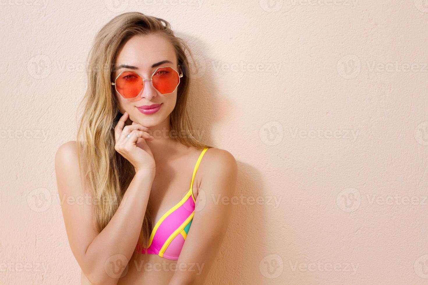 cerca de una chica feliz con gafas de sol rosas aisladas. vacaciones de verano y fin de semana divertido. concepto de verano. mujer joven sonriente en traje de baño de moda. enfoque selectivo. traje de playa de verano. copie el espacio foto