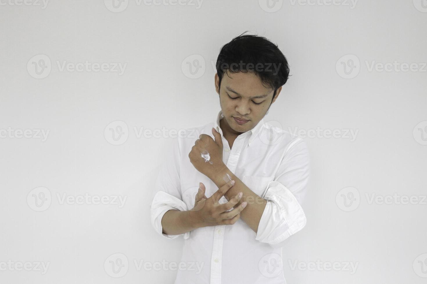 Un joven apuesto asiático aplicando crema en su mano con cara sonriente, concepto de cuidado de la piel, aislado de fondo blanco foto