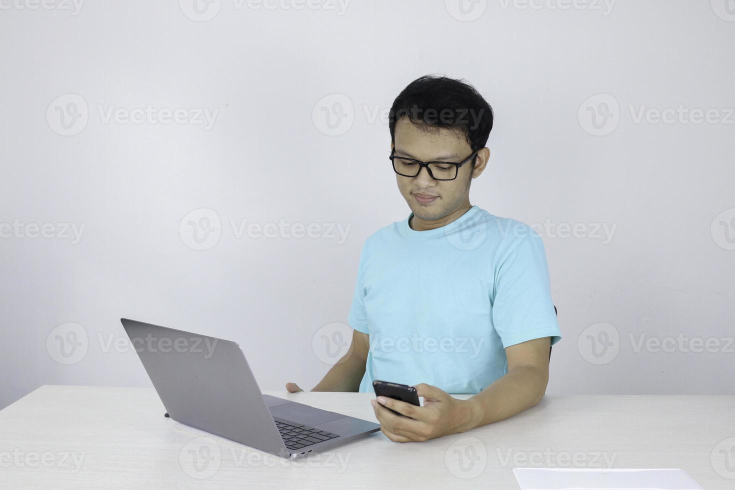 Young Asian Man is serious and focus when working on a phone and laptop on the table. Indonesian man wearing blue shirt. photo