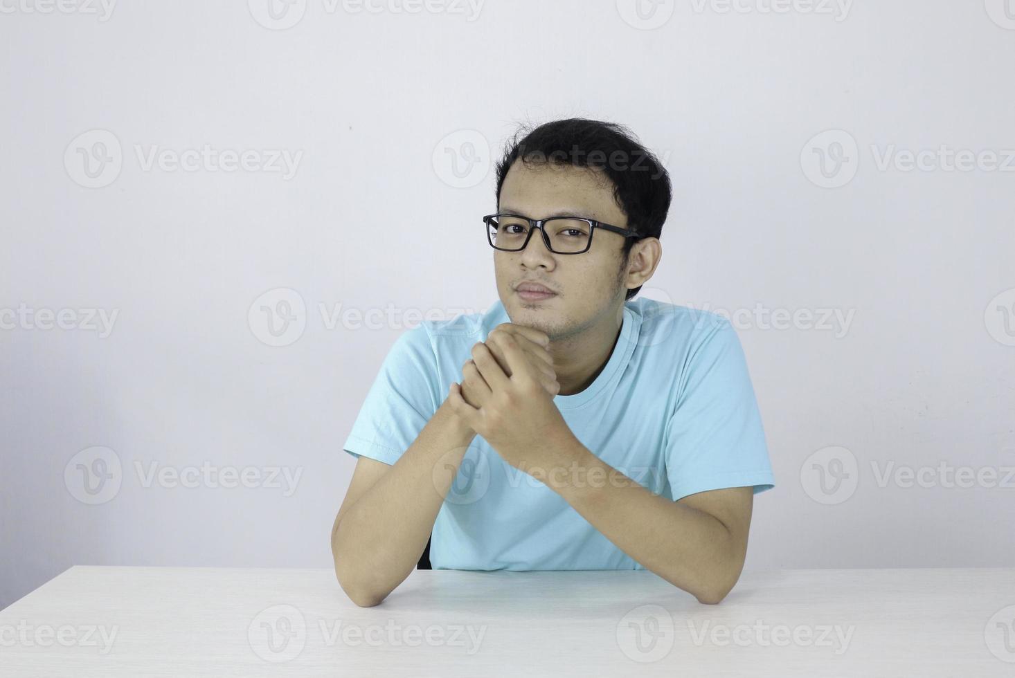 Young Asian man wear blue shirt and glasses with serious face looking on the camera and hand hold chin on the table. photo