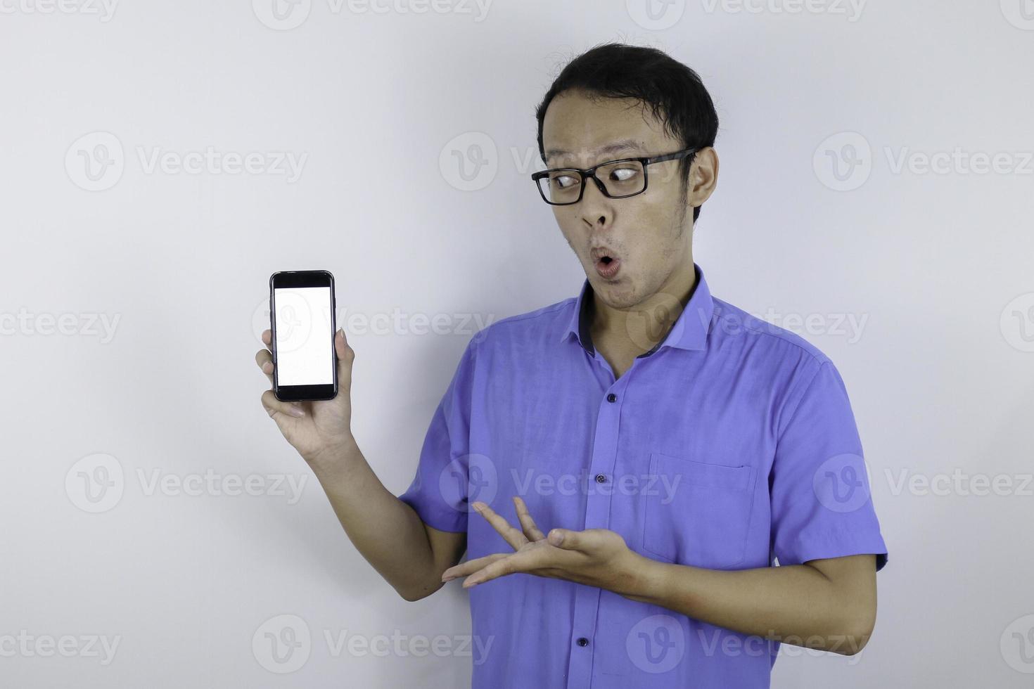 Young Asian man wear blue shirt is standing and shock with pointing on white blank space on smartphone screen on white background. photo
