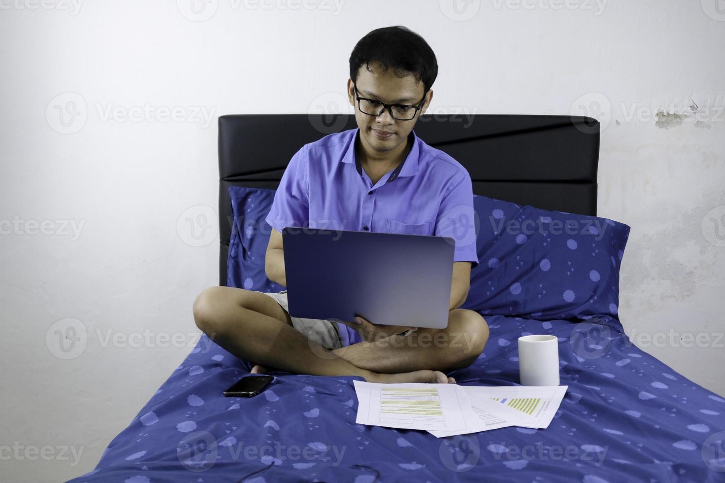 el joven asiático sonríe y disfruta cuando trabaja en casa. un hombre sentado en la cama y trabajando con una computadora portátil. trabajo desde el concepto de hogar. foto