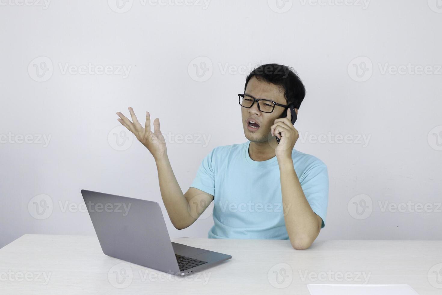 Wow face of Young Asian man shocked what he see in laptop when calling on phone. Indonesian man wearing blue shirt. photo
