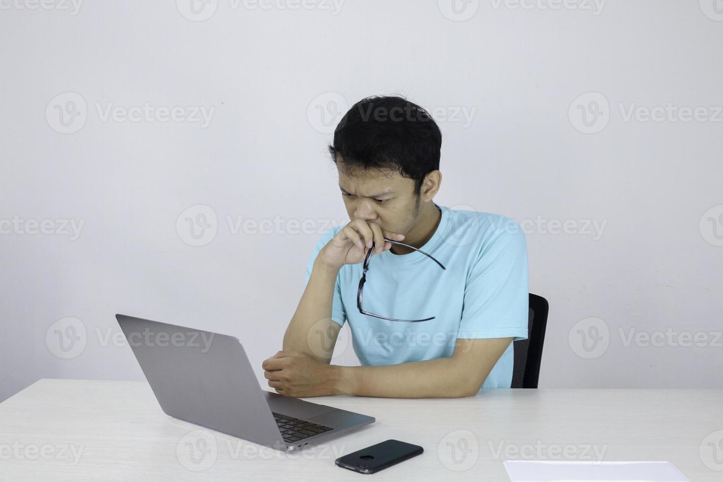 el joven asiático es serio y se enfoca cuando trabaja en una computadora portátil en la mesa. hombre indonesio con camisa azul. foto