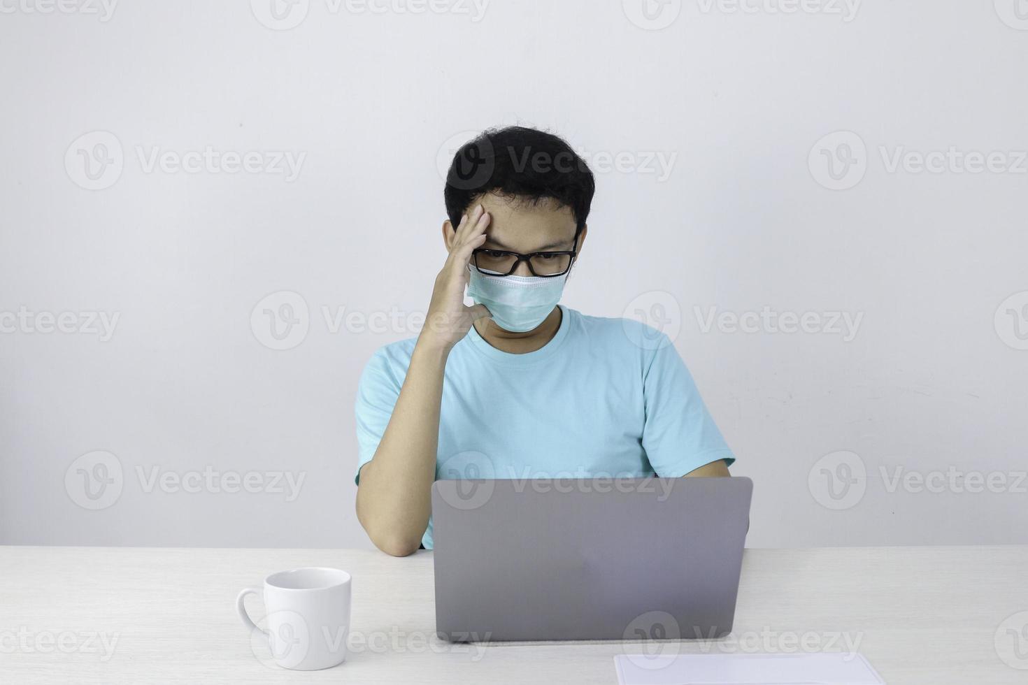 el joven asiático que usa una máscara médica no se siente saludable, está cansado y se confunde con el trabajo en una computadora portátil. foto