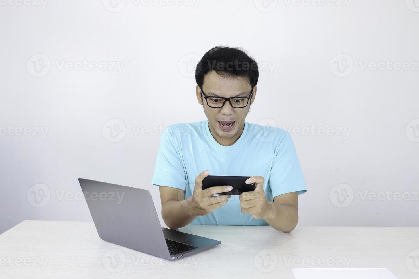 un joven asiático enojado se enoja con el teléfono inteligente cuando juega en el trabajo. hombre indonesio con camisa azul. foto