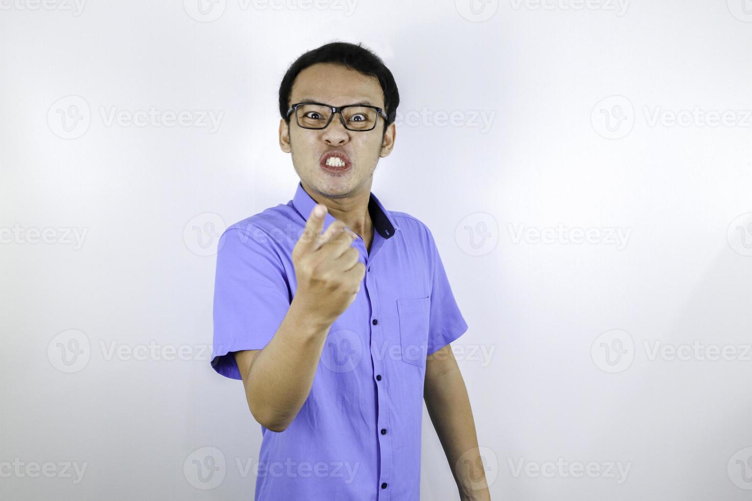 El joven asiático que usa camisa azul es una cara graciosa y enojada con gritos y señalando con el dedo a la cámara aislada sobre el fondo blanco. foto