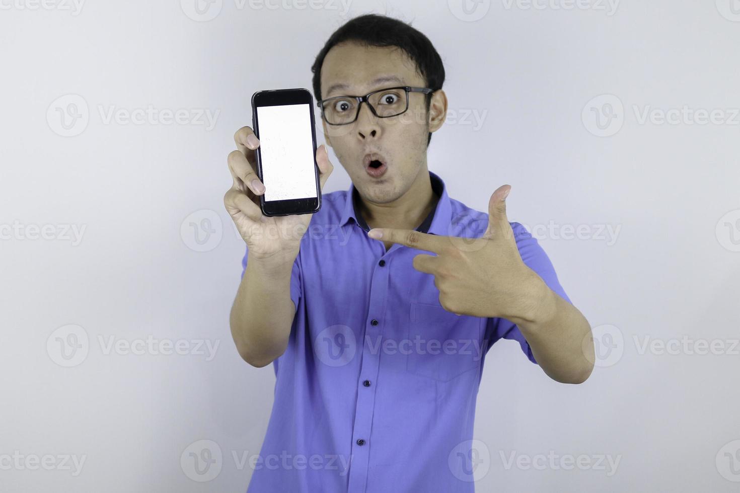 Young Asian man wear blue shirt is standing and shock with pointing on white blank space on smartphone screen on white background. photo