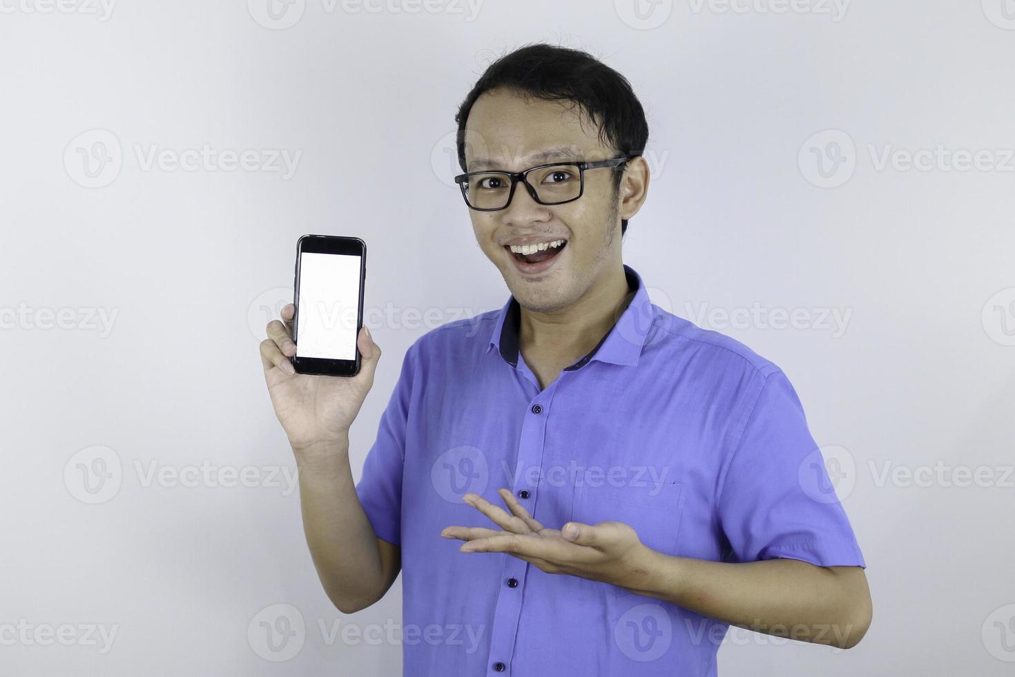 un joven asiático con camisa azul está de pie y sonriendo señalando un espacio en blanco en la pantalla del teléfono inteligente con fondo blanco. foto