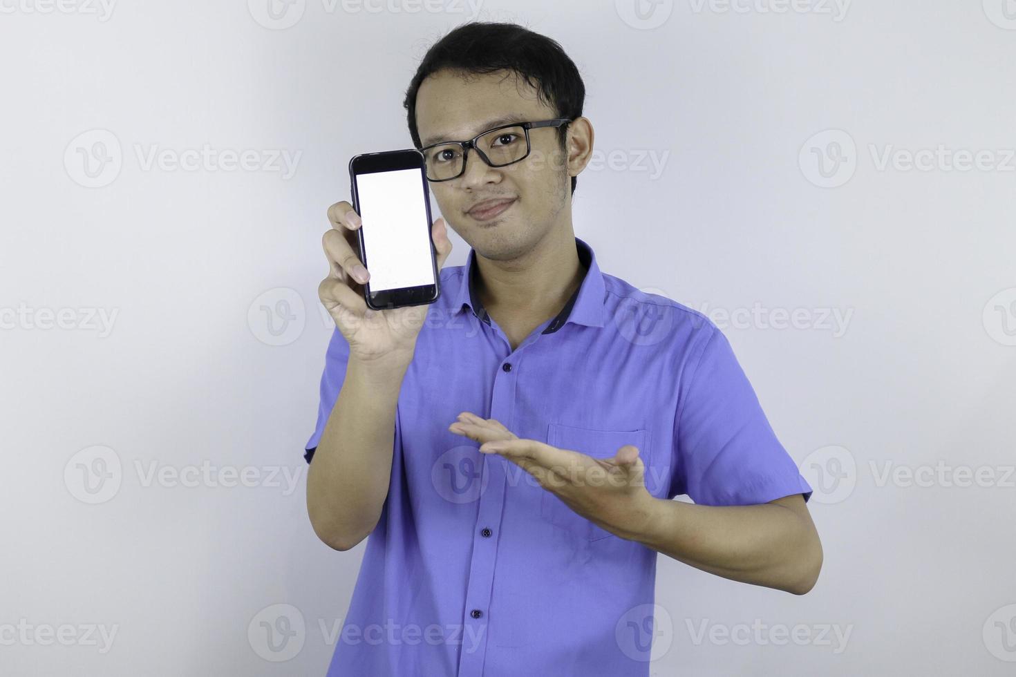 Young Asian man wear blue shirt is standing and smiling pointing on white blank space on smartphone screen on white background. photo