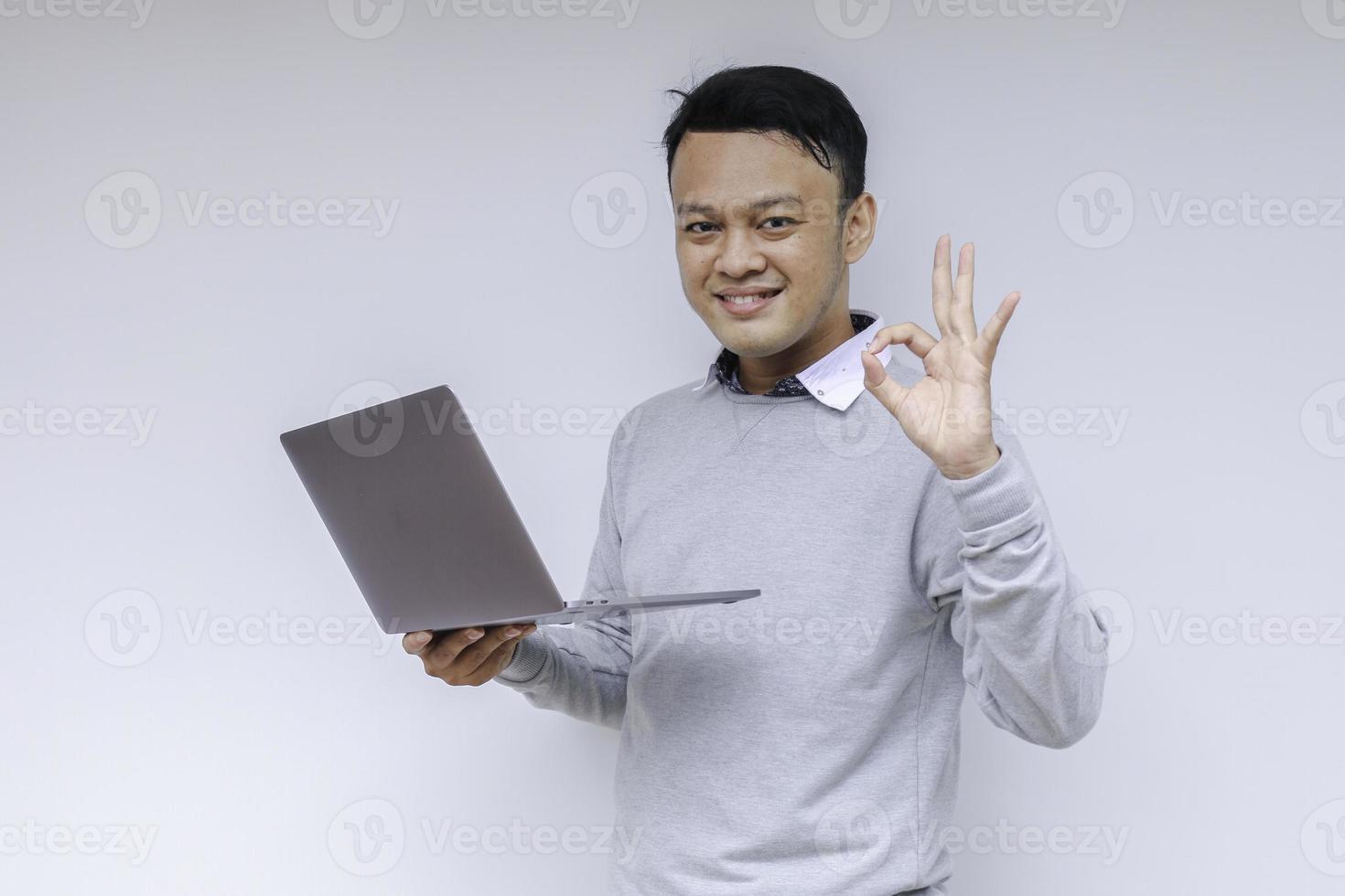 Young Asian man feeling happy and smile when standing and work laptop. Indonesian man wearing grey shirt photo