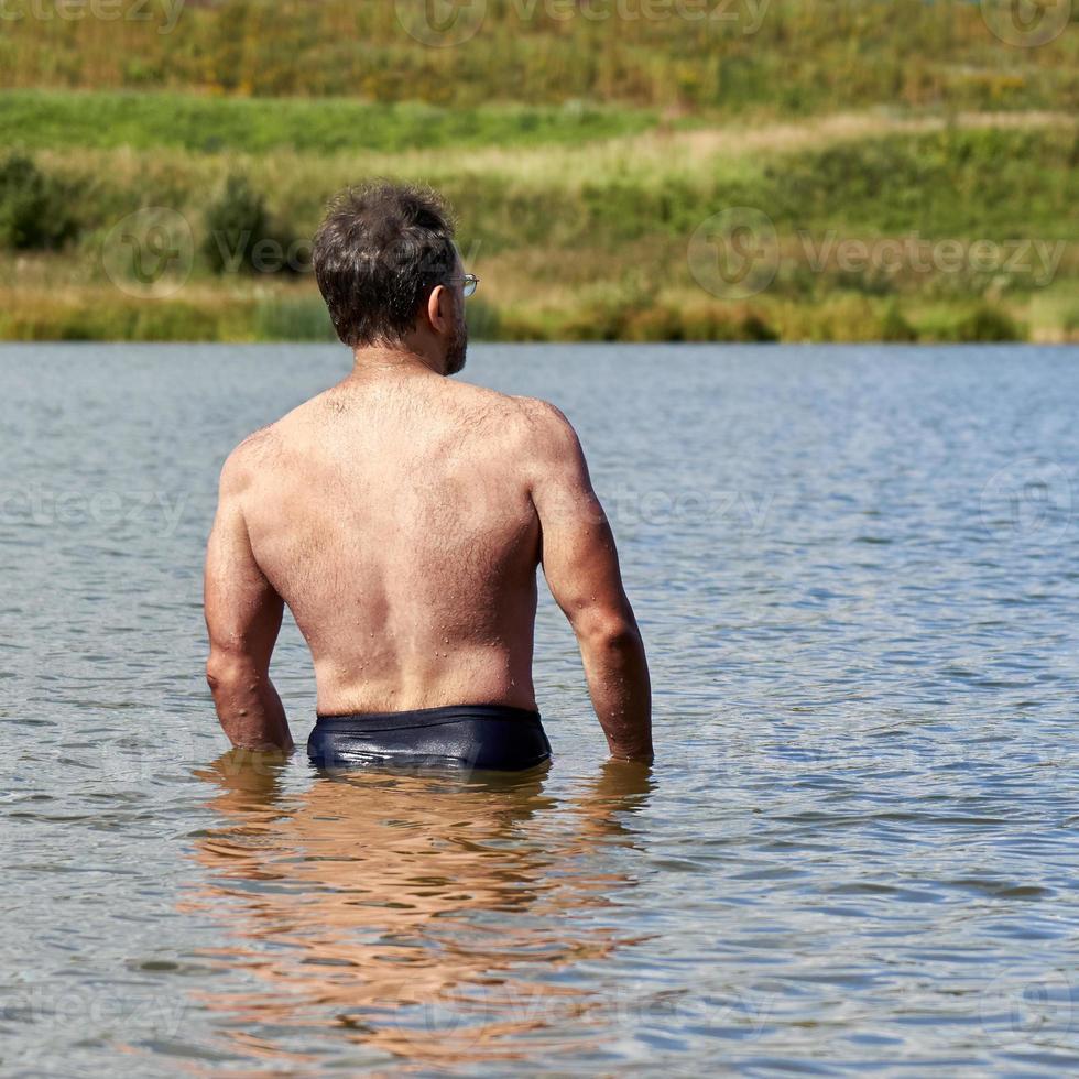 Muscular middle-aged man is bathing in the small suburban lake in summer. Back view photo