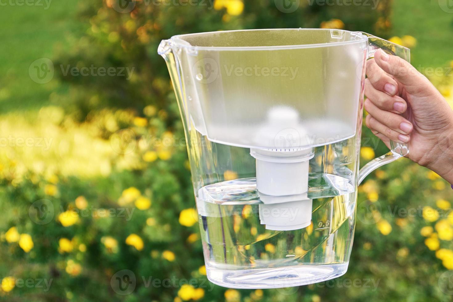 Girl's hand holds a water filter jug in the sunny summer garden with yellow flowers on the background photo