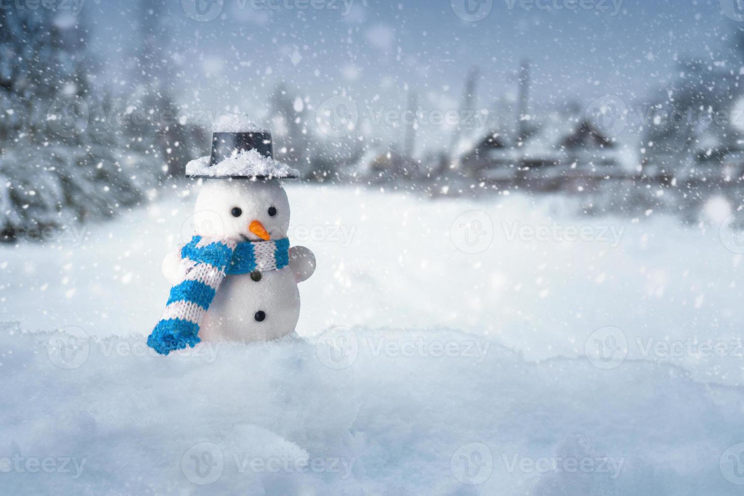 Snowman stands in a snowdrift with a village on the background in snowy evening photo
