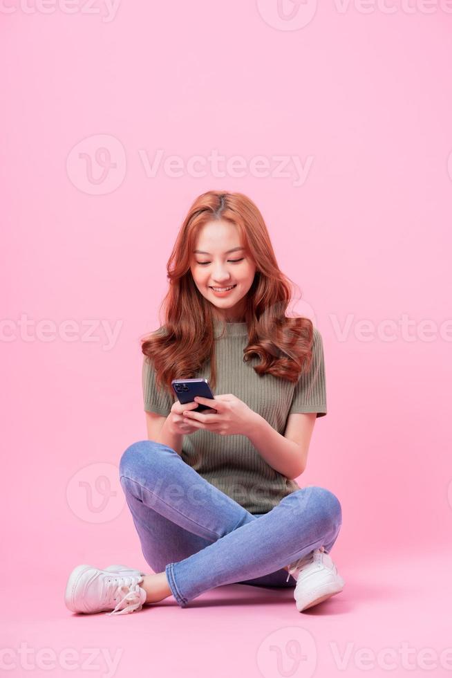 Young Asian woman sitting and using smartphone on pink background photo