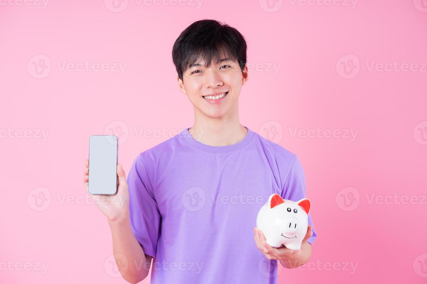 Young Asian man holding piggy bank on pink background photo