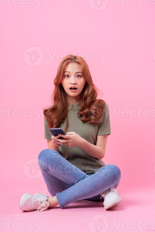 joven asiática sentada y usando un teléfono inteligente con fondo rosa foto