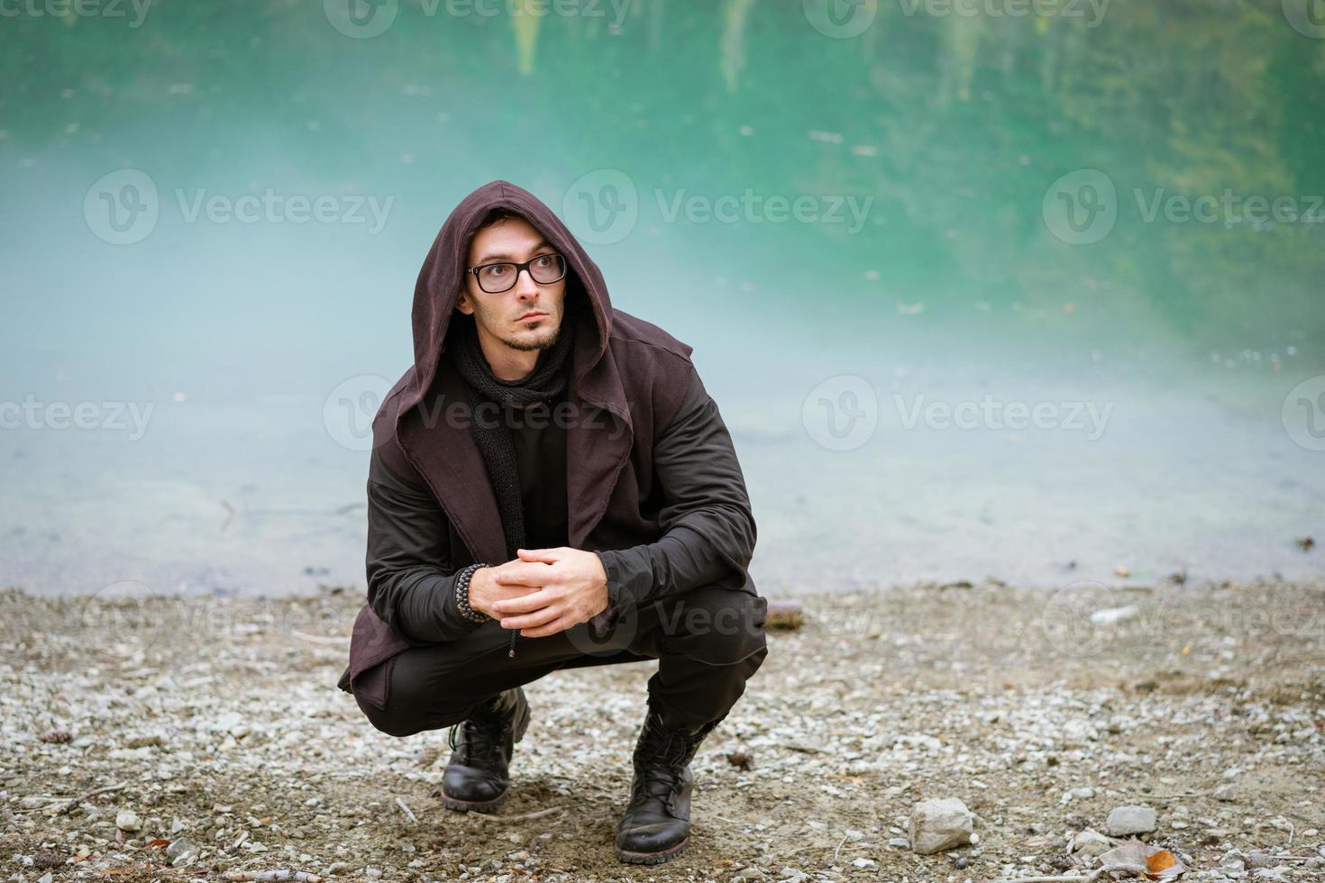 Bearded man in casual clothes with hood and glasses stands on shore photo