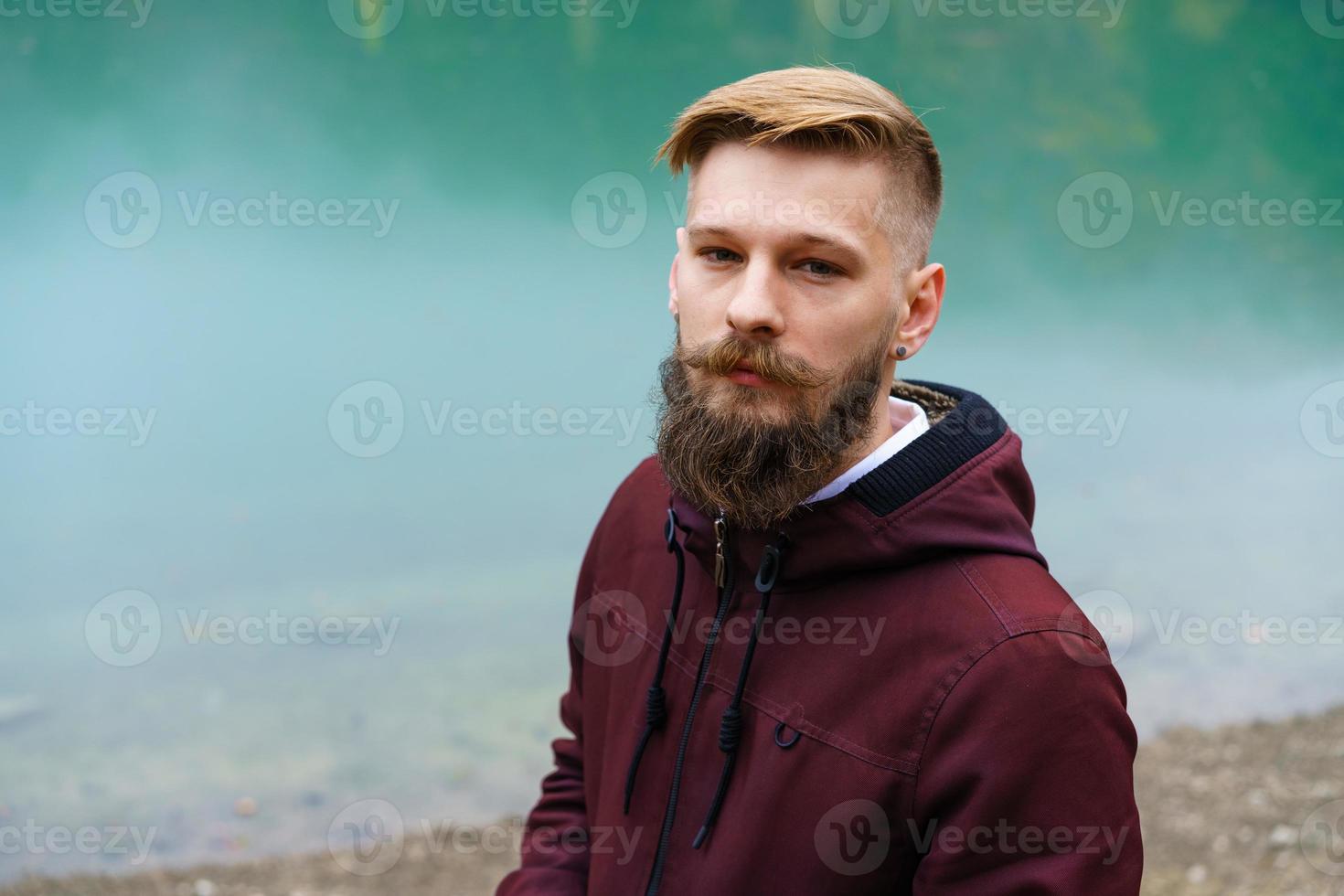 Young bearded man stood alone by lake and looked aside thoughtfully smoking photo