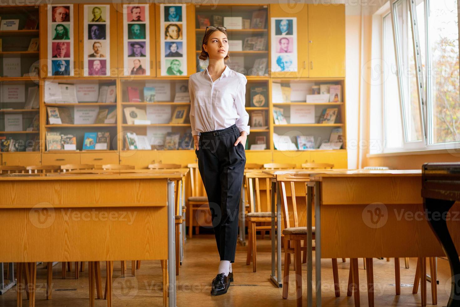 una hermosa chica con una camisa blanca se para en el salón de clases entre los escritorios foto